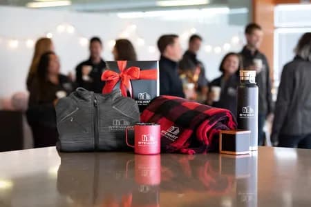 Branded company gifts including custom logo jacket, mug, blanket, and bottle displayed on a table with coworkers in the background.