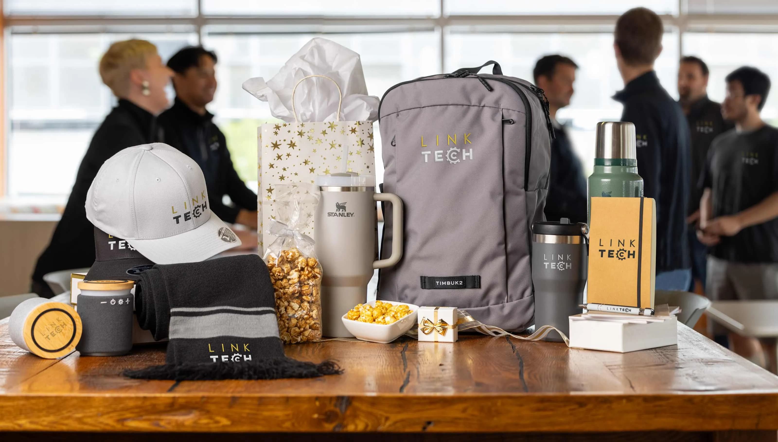 Branded company merchandise including custom logo hats, mugs, backpack, and notebooks displayed on a table with coworkers in the background.