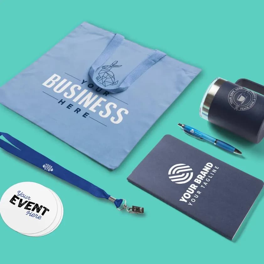 A laydown of branded promotional products including a tote bag, lanyard, notebook, pen, stickers, and insulated mug.